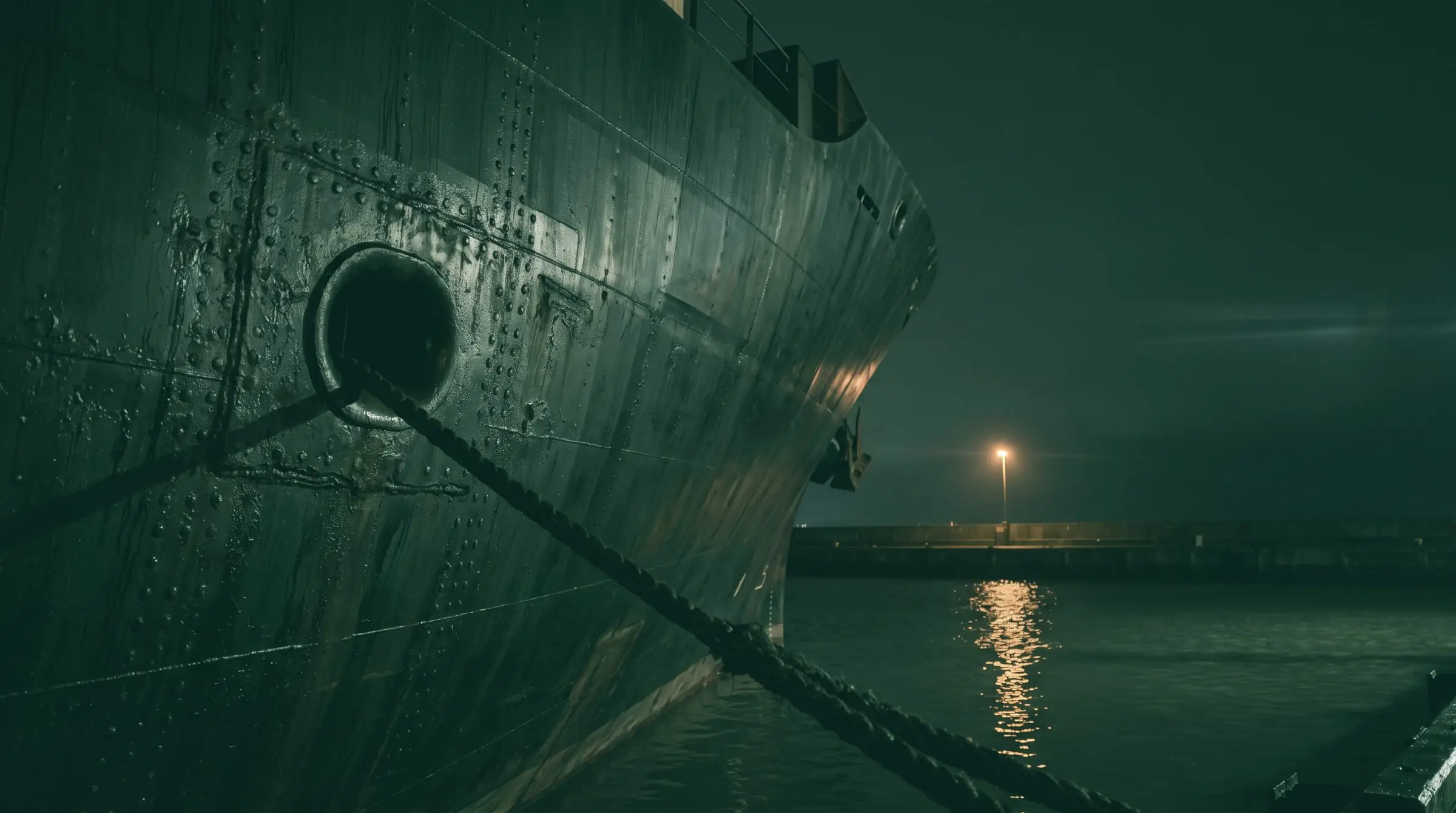 The steel hull of a cargo ship at berth at night, a mooring line taut across dark water.